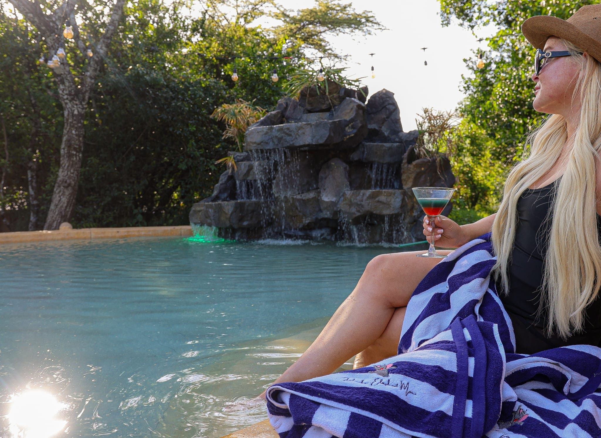 Guest with a cocktail and striped towel at the pool's edge, facing a rock waterfall