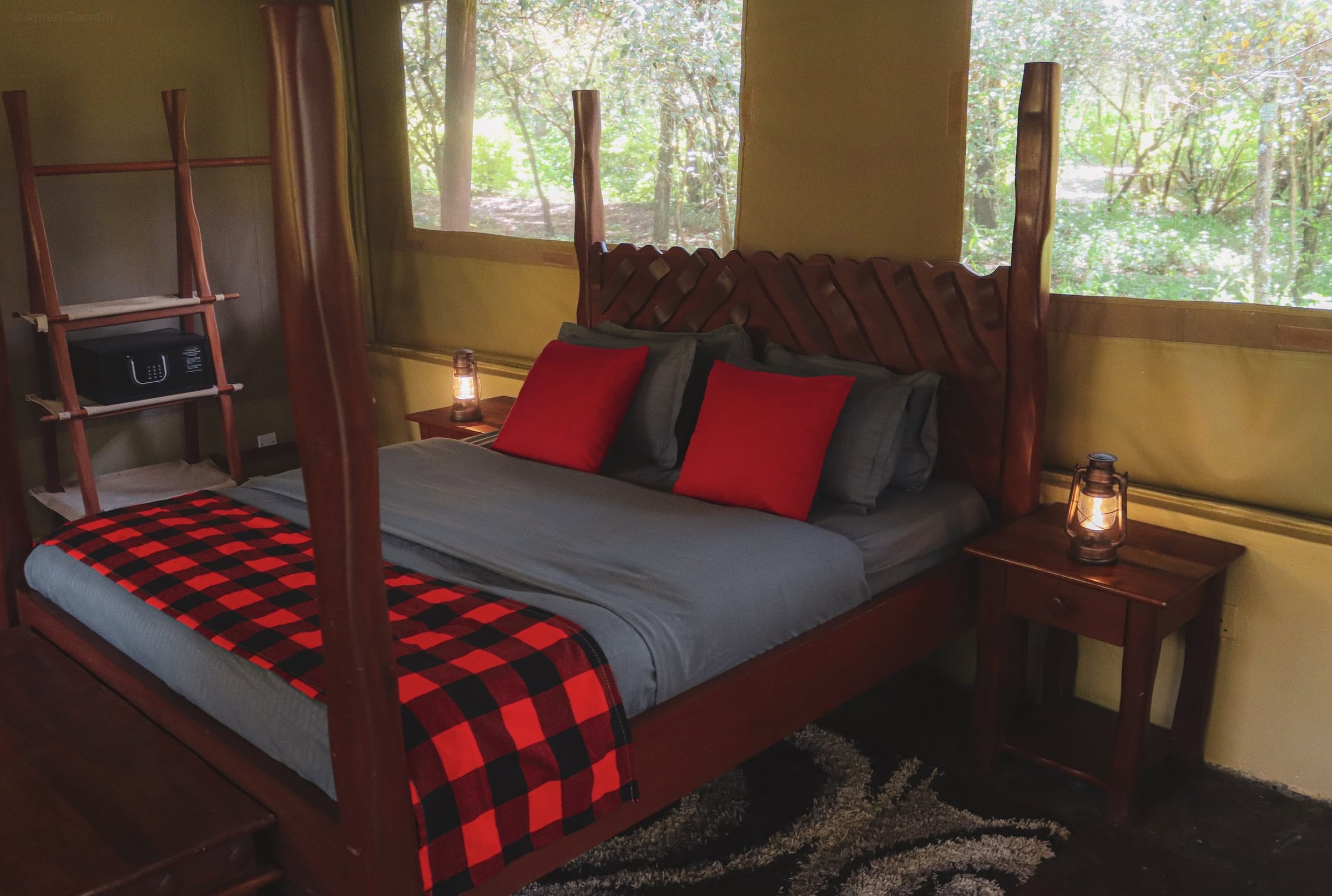 Double Tent: hand-carved four-poster bed with grey linen, a red Maasai-check blanket and glowing lanterns, forest through the windows