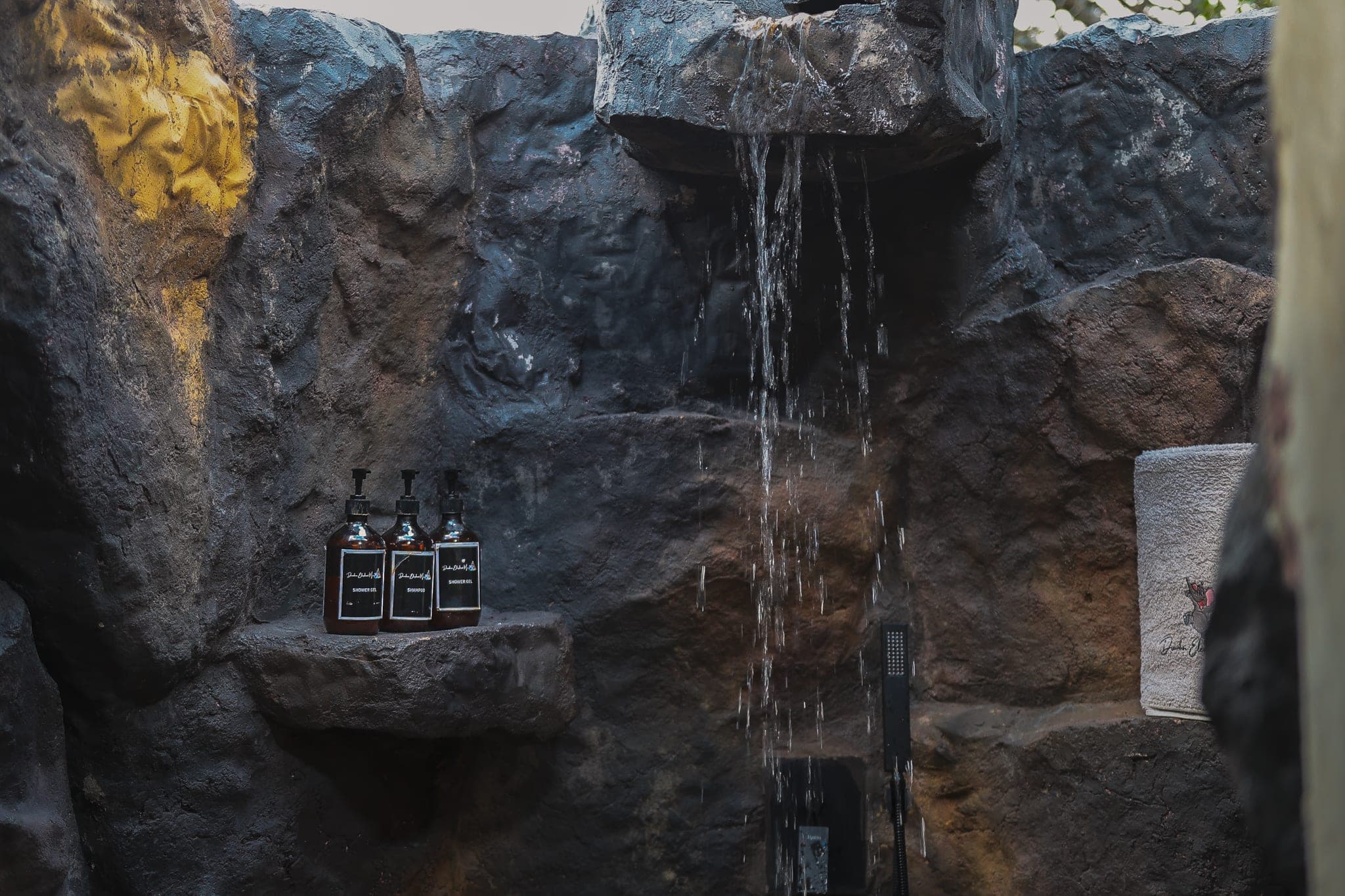 Outdoor rock shower with water pouring from a stone ledge, amber toiletry bottles and a folded towel