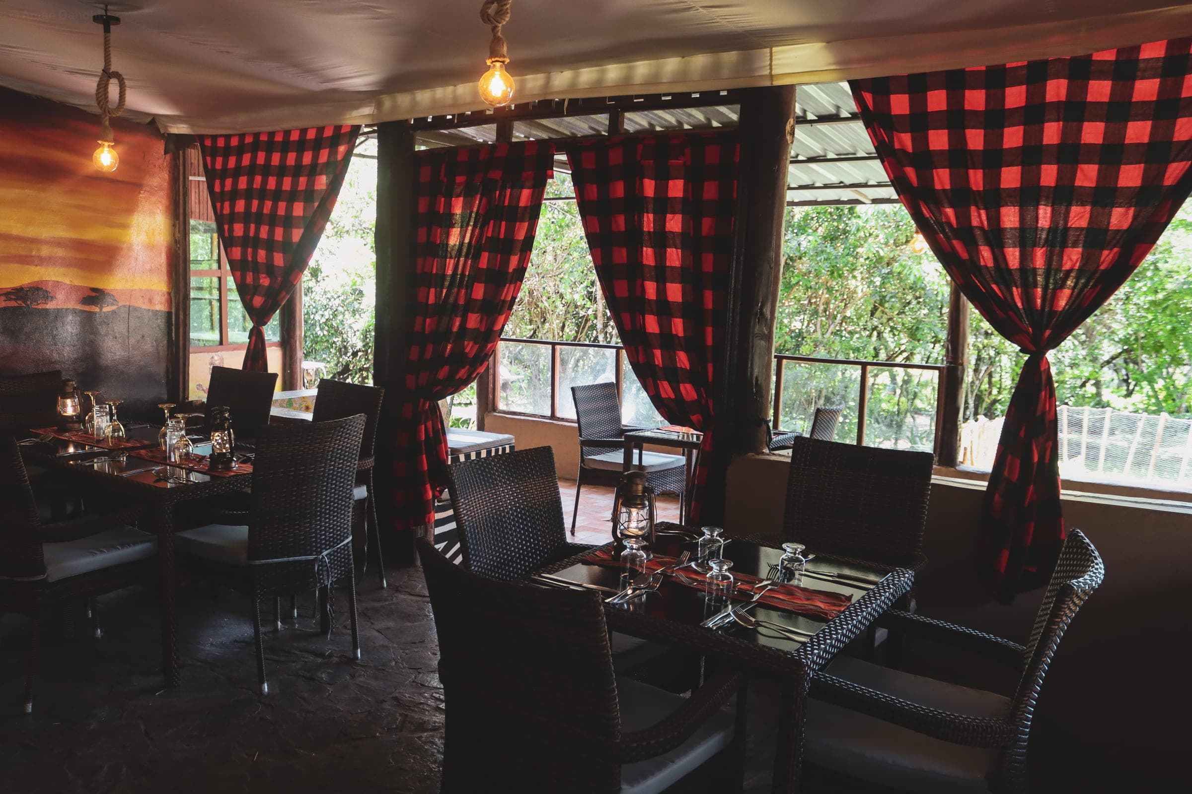 Dining tent with red check curtains, rattan chairs and lantern-set tables, open to the trees