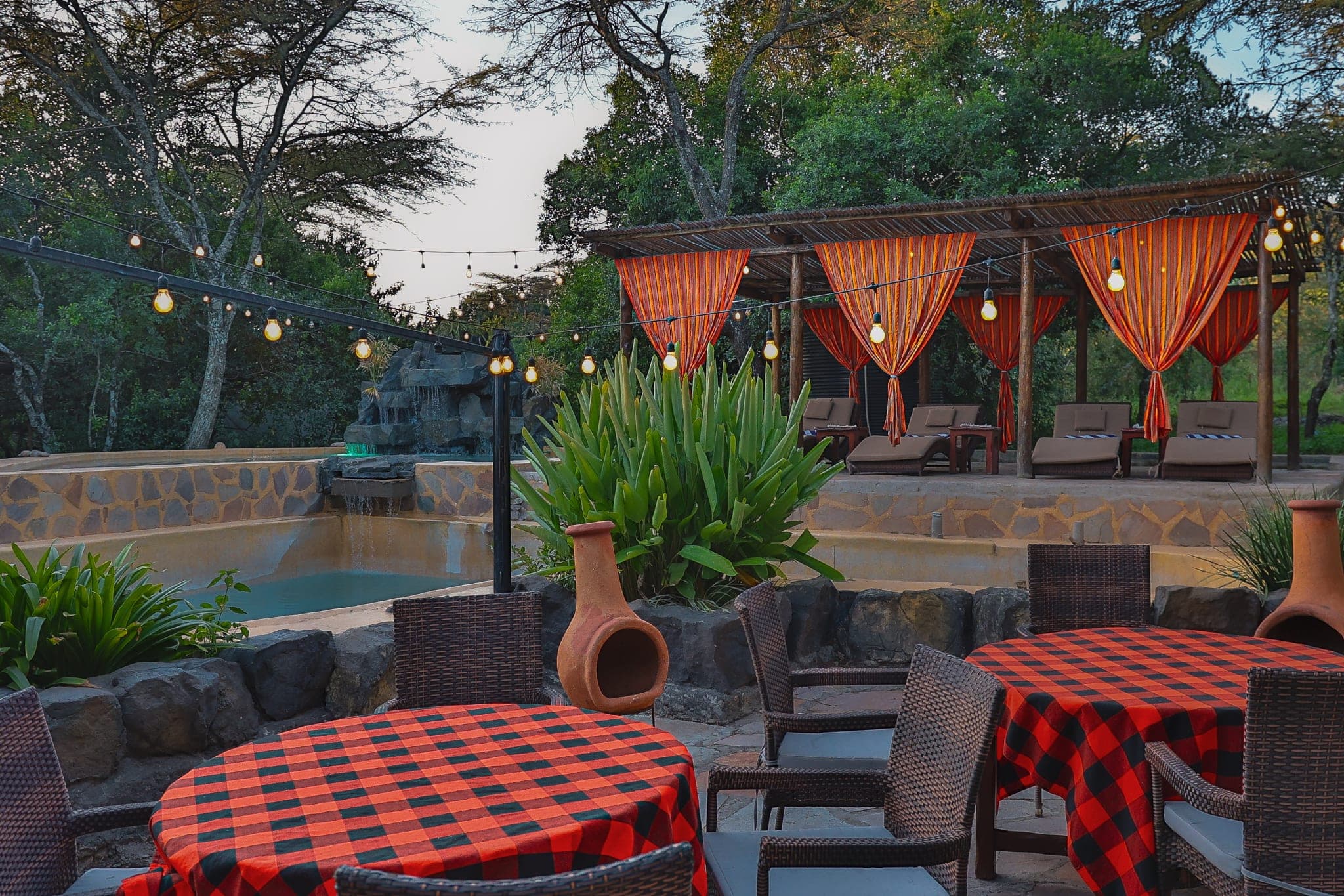 Poolside tables with red check cloths under festoon lights at dusk, curtained cabanas behind