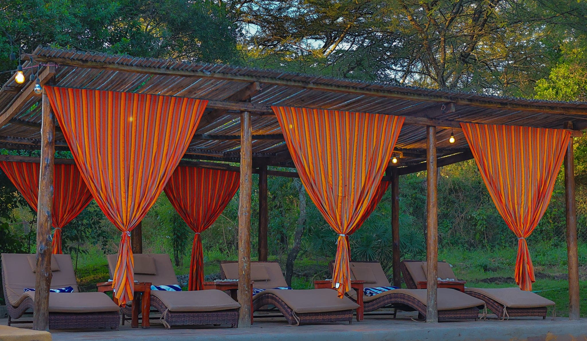 Loungers under a thatched pergola hung with orange striped curtains, trees behind