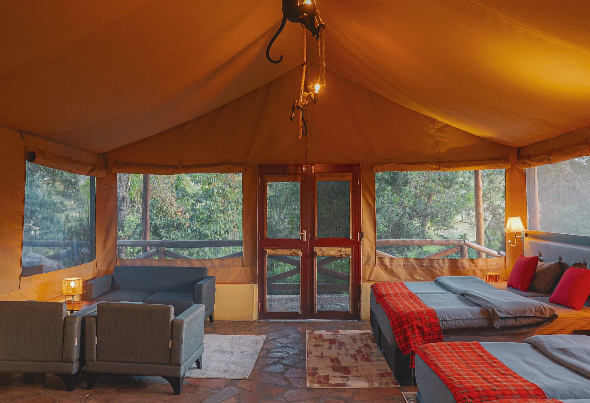 Lamplit family tent with a sofa and armchairs, glazed doors to the deck and twin beds with red check throws