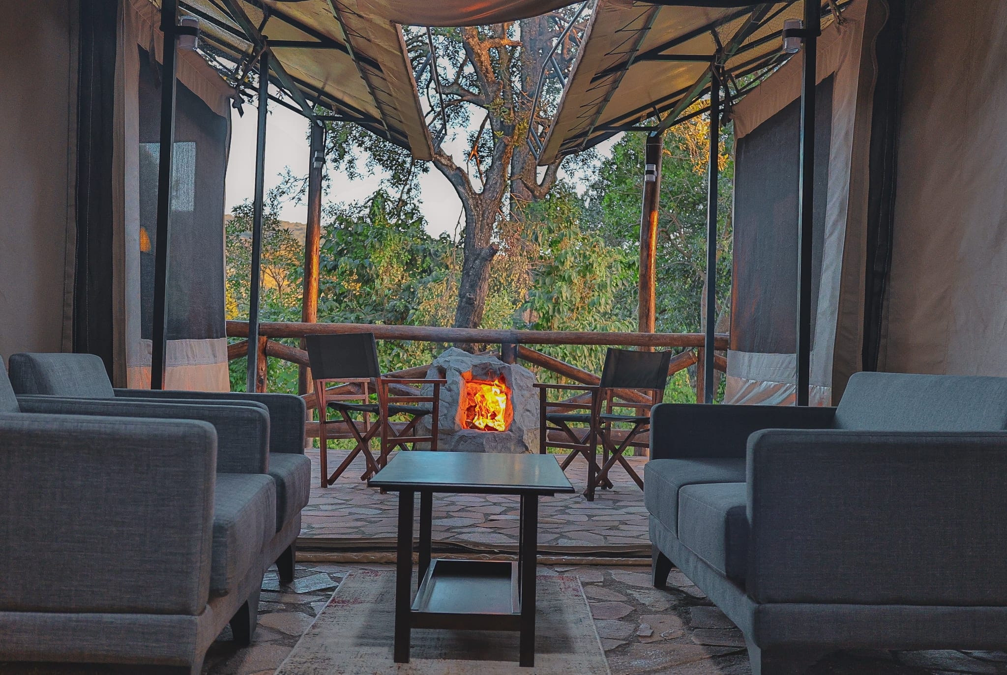 Tent lounge with grey sofas looking out to a lit stone fire pit on the deck and forest trees at golden hour